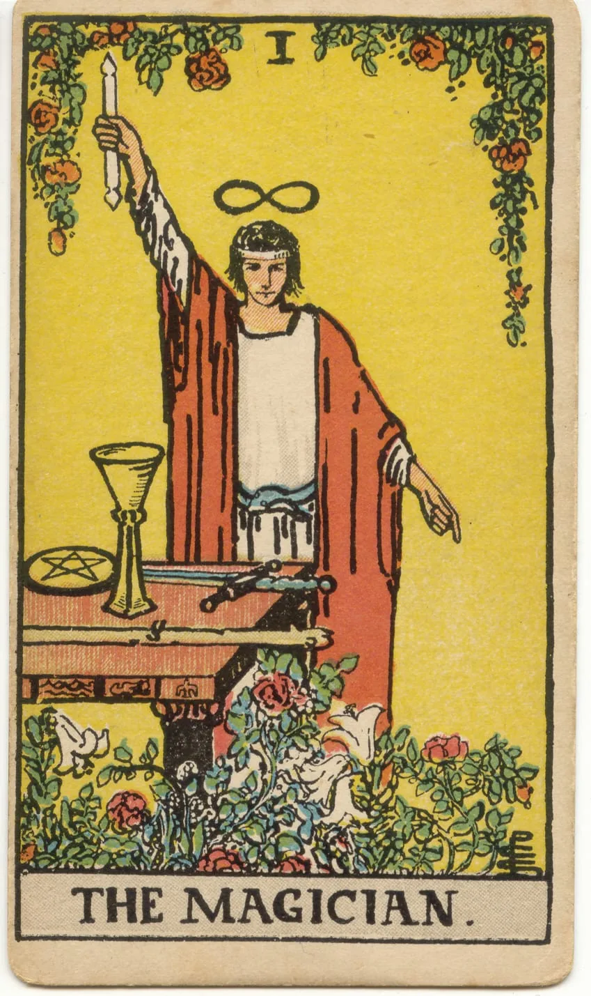 O Mago diante de uma mesa com taça, espada, bastão e pentáculo, com uma varinha erguida e a outra mão apontando para a terra.