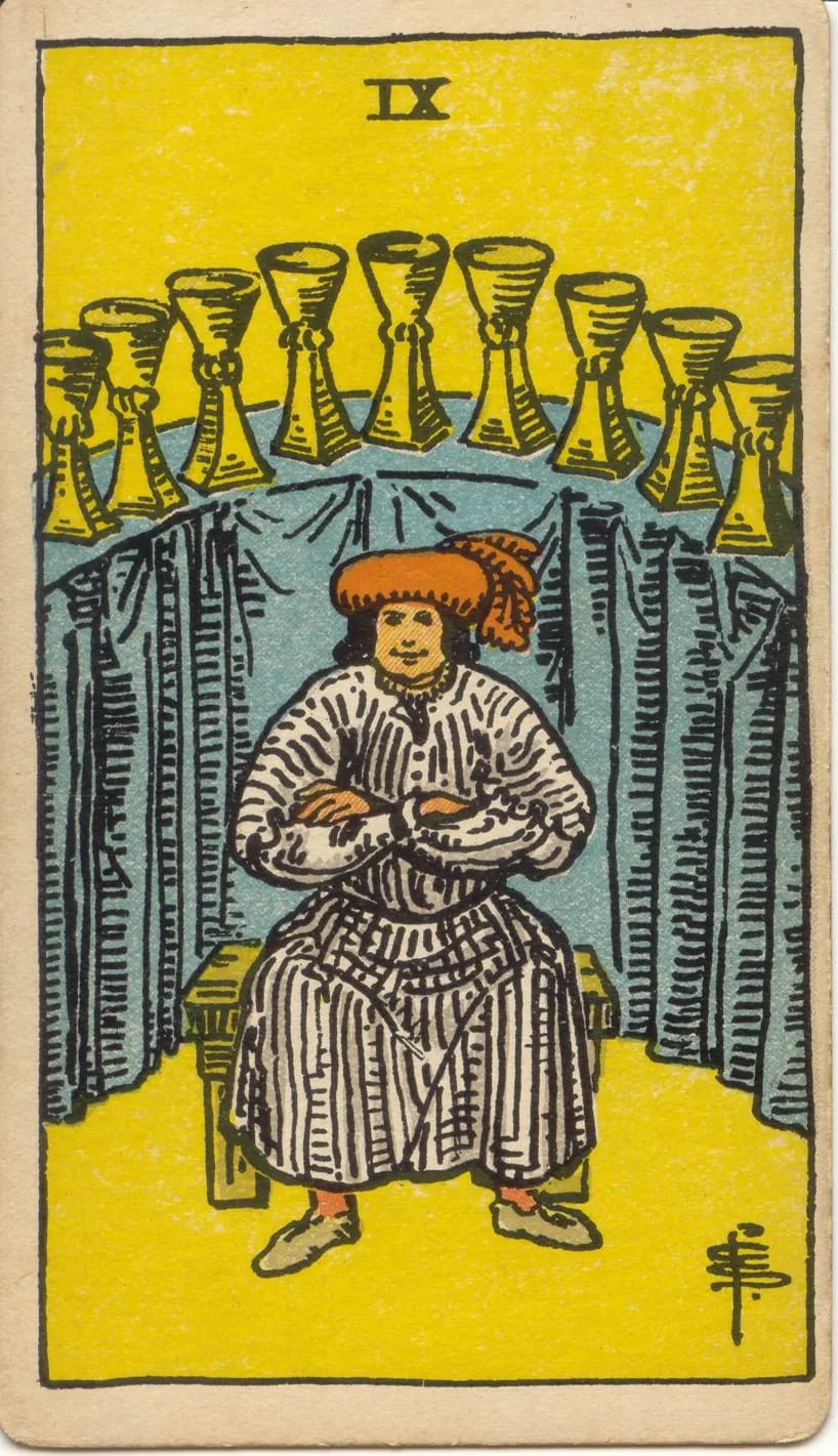 Pessoa sentada de frente, com braços cruzados e gorro vermelho, diante de nove taças sobre um balcão coberto com tecido azul.