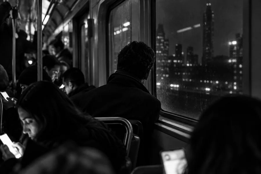 Passageiros em vagão de metrô olham para celulares, enquanto um homem de costas observa a cidade pela janela à noite.